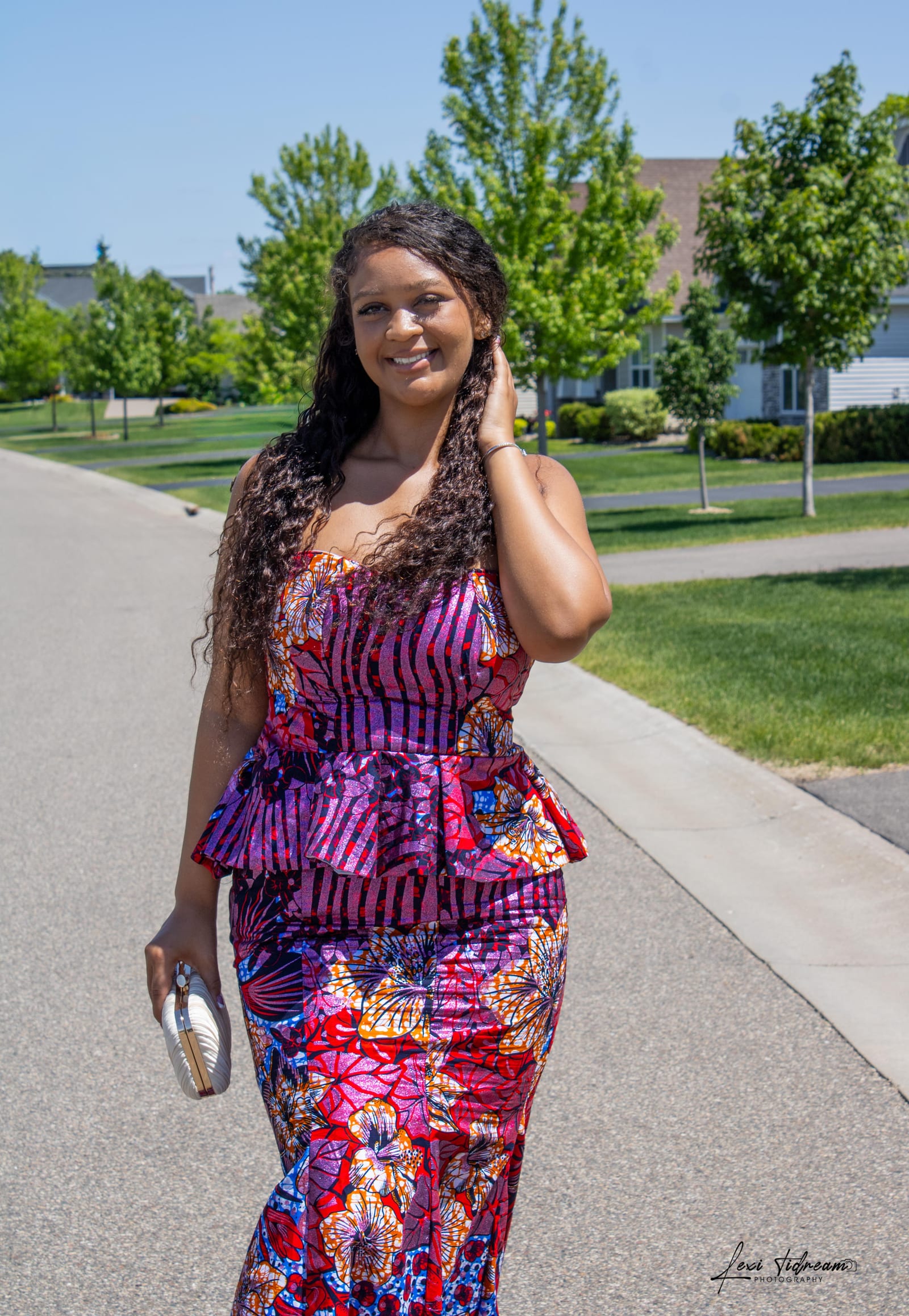 Woman in a vibrant magenta and purple patterned dress smiling on a neighborhood street