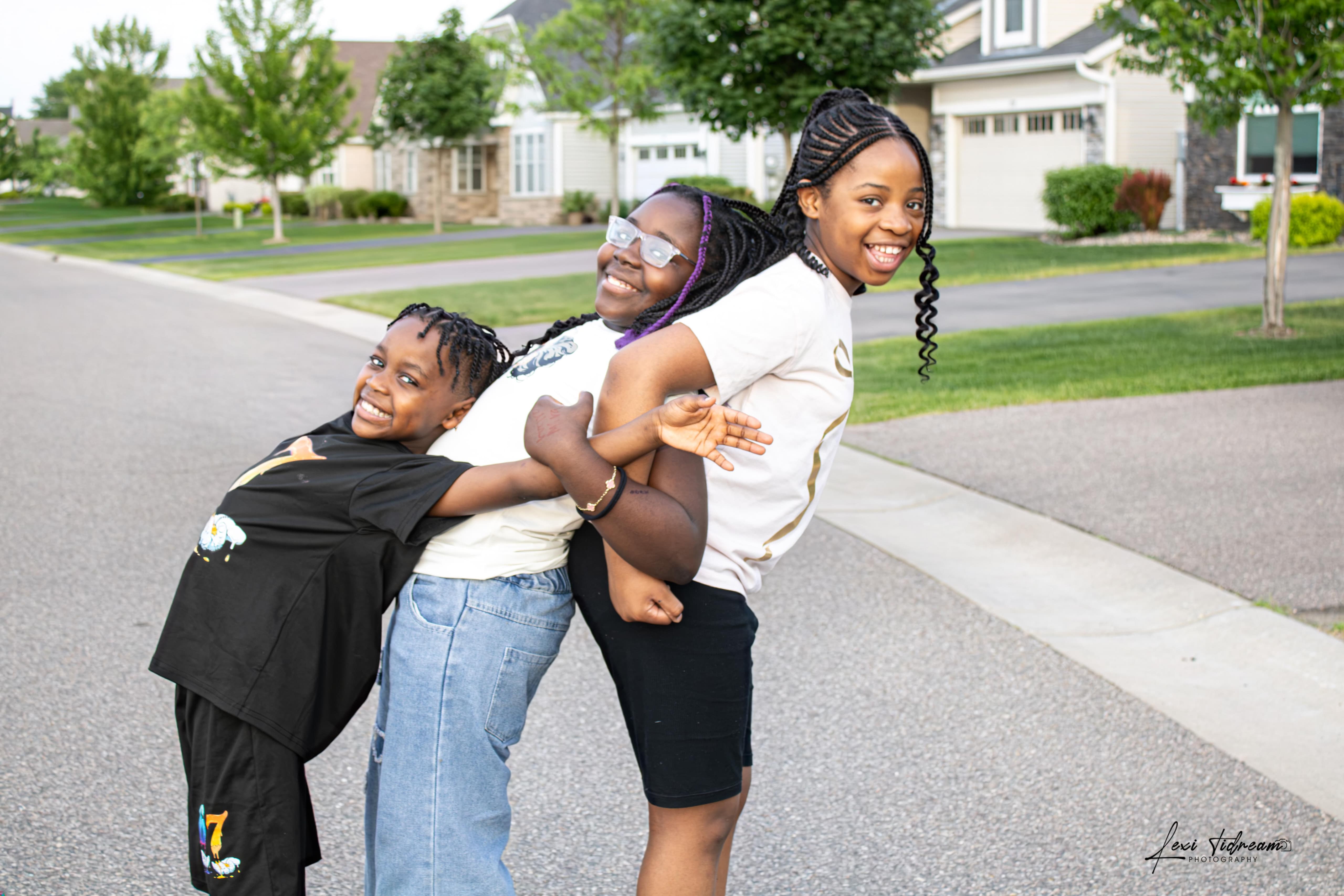 Three sisters laughing together, arms wrapped around each other, on a neighborhood street
