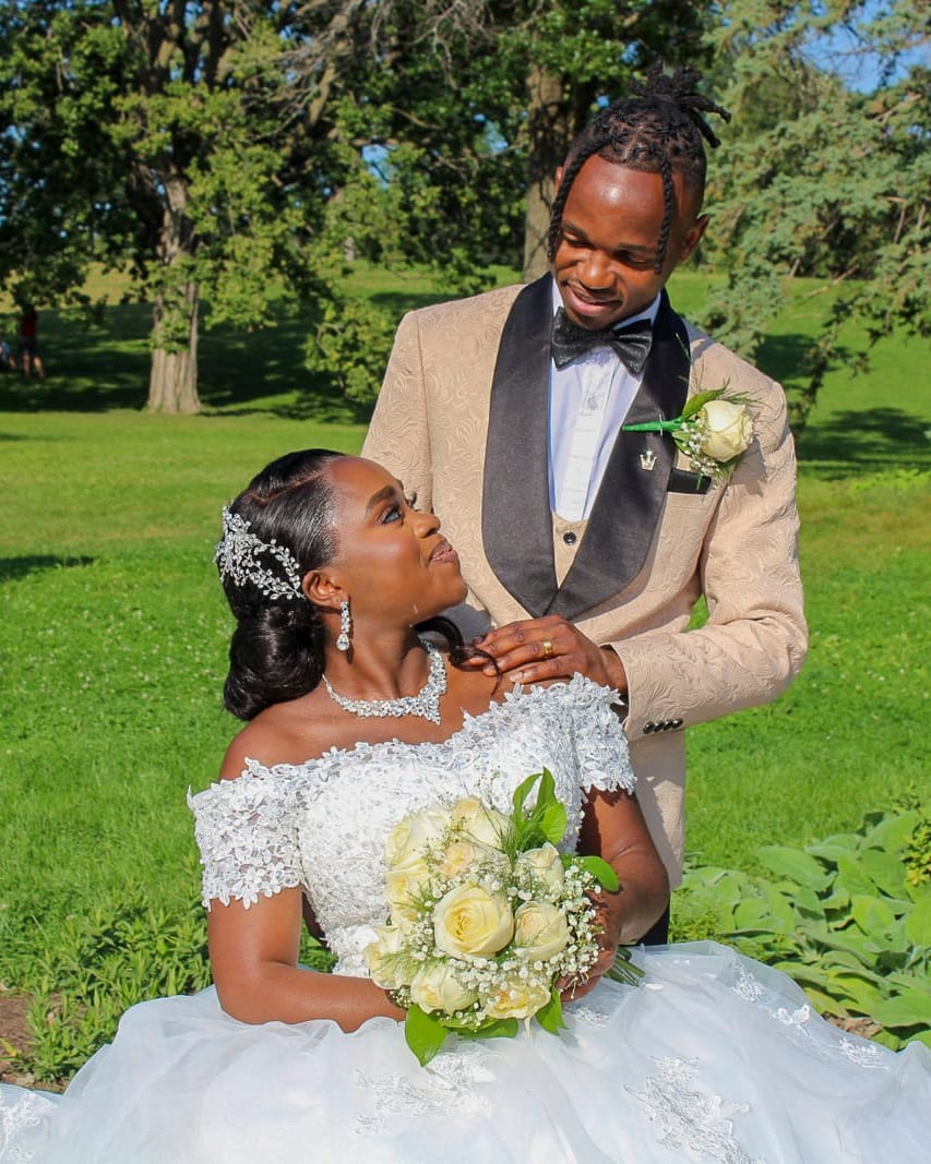 A bride seated with her groom standing behind her in a park