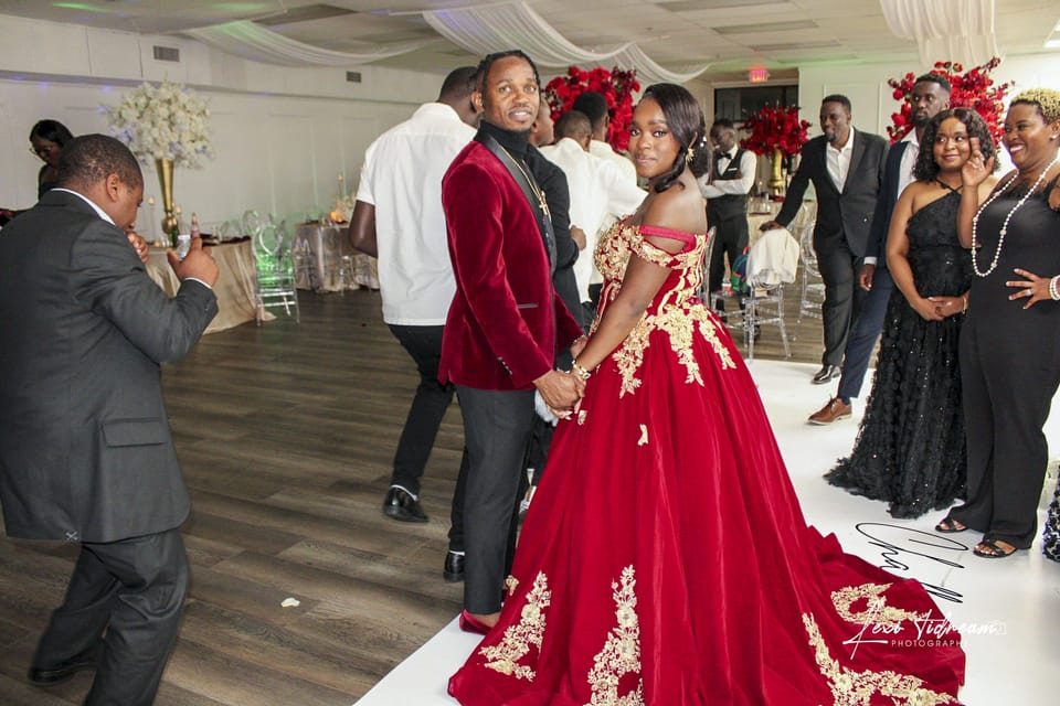 Couple at a celebration reception, one in a red and gold ball gown, surrounded by guests