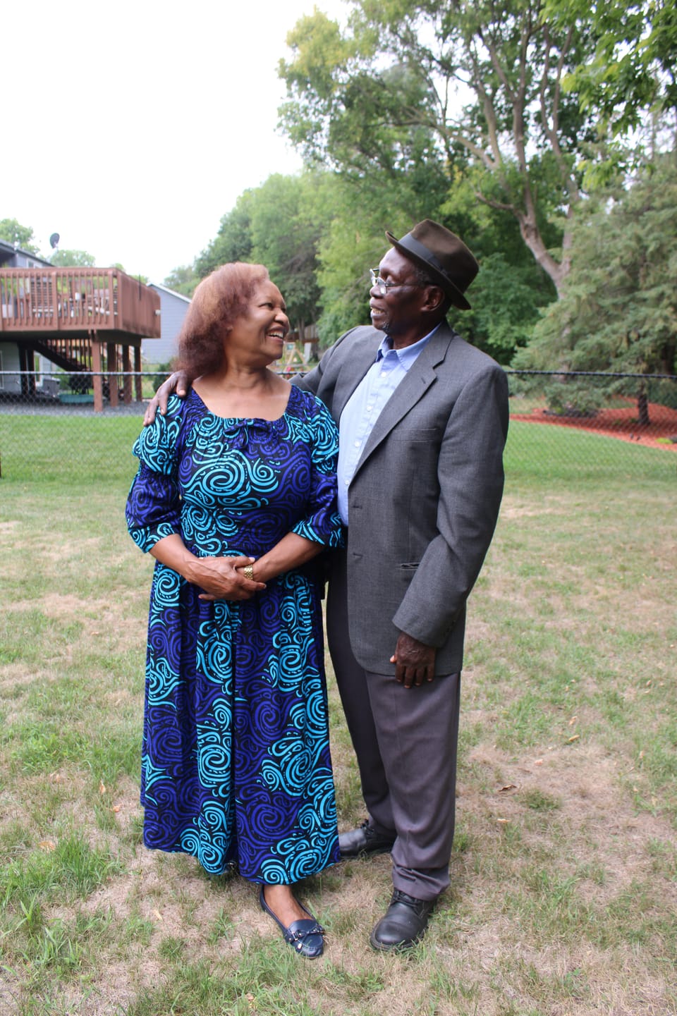 Elderly couple outdoors, one in a blue patterned dress and one in a grey suit and hat, smiling at each other