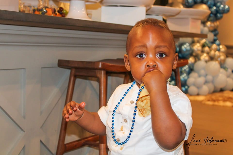 Toddler in a white outfit with blue beads at a balloon-decorated birthday party