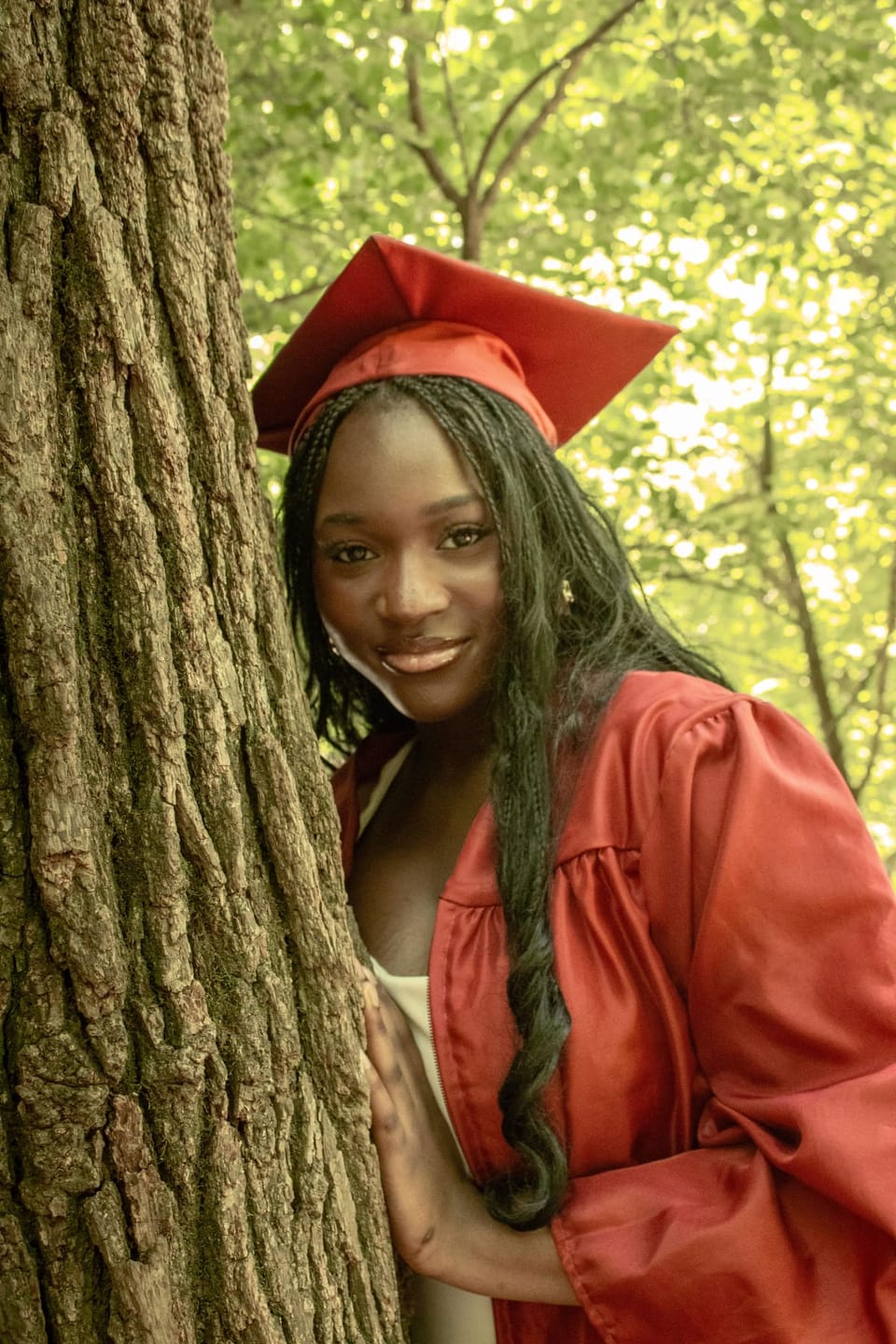 High school graduate in a red cap and gown smiling beside a tree