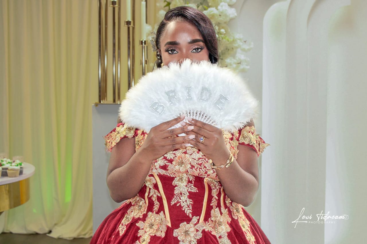 A bride in a red and gold gown holding a white feather fan lettered BRIDE