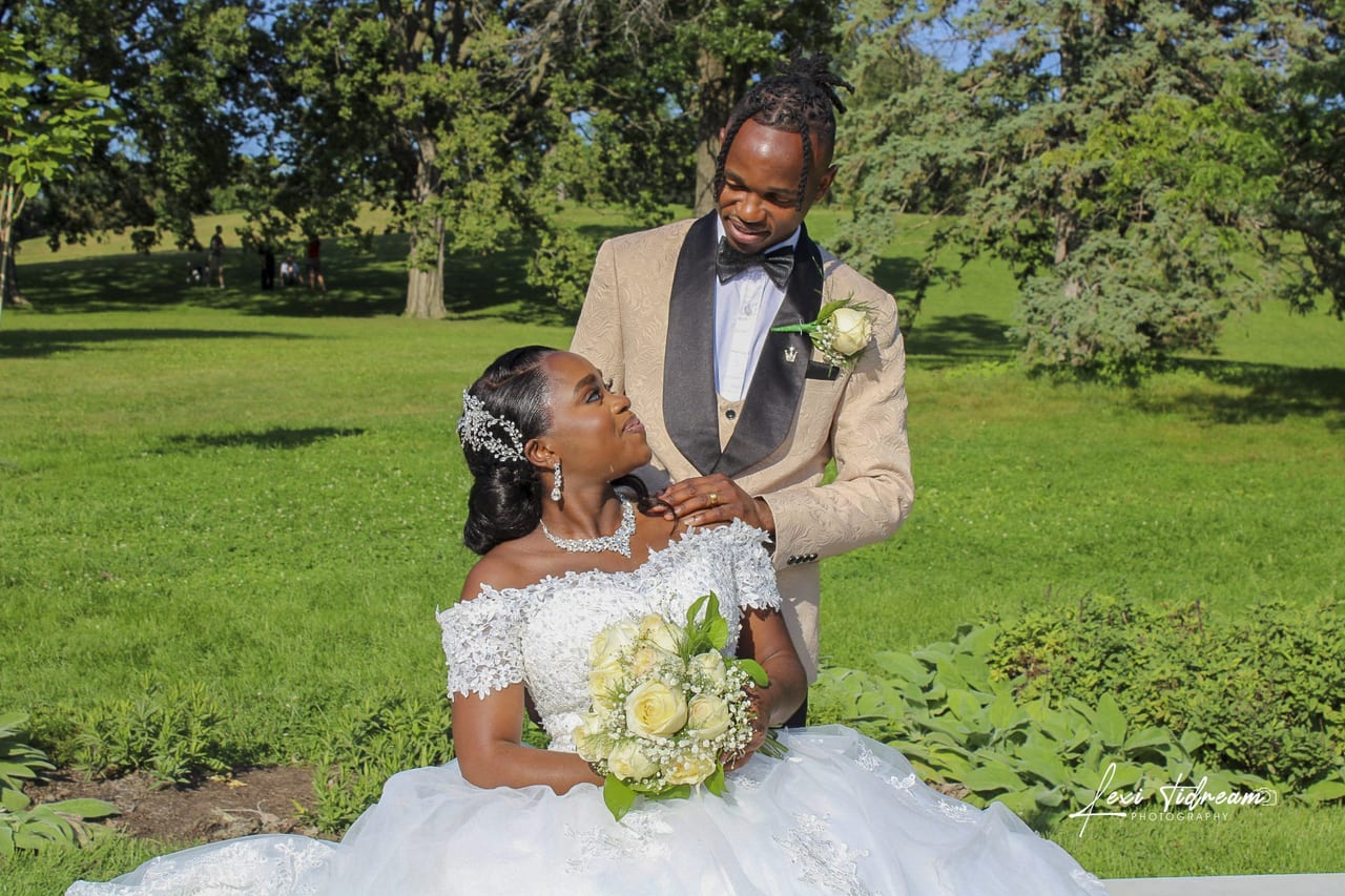A bride seated in a white gown with her groom standing behind her in a park